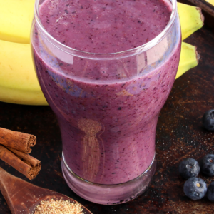 blueberry smoothie with a banana in the back and cinnamon and flax seeds next to it